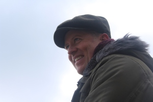 Side profile of a man with a flat cap, smiling and looking into the distance. He is wearing a burgundy scarf and an olive-green coat with a fluffy hood.